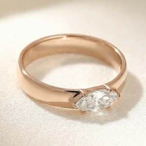 May include: A rose gold ring with a marquise-cut diamond center stone. The band is smooth and polished, and the diamond is set in a bezel setting. The ring is displayed on a white, textured surface.