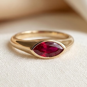 May include: A gold ring with a marquise-cut, deep red gemstone. The ring has a smooth, polished band and a bezel setting that securely holds the gemstone. The ring is displayed on a white fabric surface.