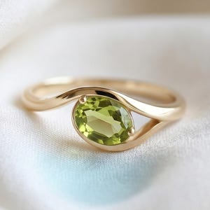 Könnte beinhalten: Ein goldener Ring mit einem einzelnen oval geschliffenen, hellgrünen Peridot-Edelstein. Die Ringschiene hat eine glatte, polierte Oberfläche und schwingt sich um den Stein, wodurch ein offenes, asymmetrisches Design entsteht.