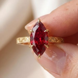 May include: A gold ring featuring a marquise-cut, deep red gemstone. The ring's band has an ornate, engraved design. The gemstone is held in place by six prongs. The ring is held up by a person's hand.