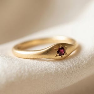 May include: A gold ring with a starburst design around a small, round, red gemstone. The ring is set on a cream-colored fabric, showcasing its polished finish and intricate details. The ring's band is smooth and rounded.