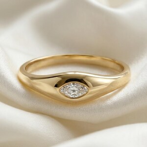 May include: A gold ring featuring a smooth, polished band with a central marquise-cut clear gemstone set horizontally. The ring has a tapered design that widens toward the center, showcasing a single faceted stone embedded within the metal surface.