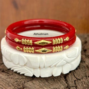 Peut inclure: Deux bracelets rouges avec des ornements dorés, posés sur un support blanc sculpté. Les bracelets ont une finition brillante et sont ornés de motifs dorés complexes. Le texte "Abhushnam" est visible.