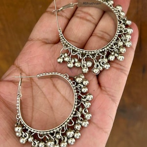May include: Silver-toned hoop earrings with dangling ball accents. The earrings have a hinged closure and are embellished with small, round beads along the bottom edge. The word "Abhushnam" is printed on the top of the earring.