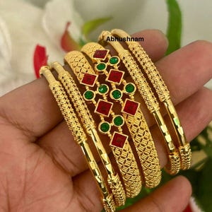 Peut inclure: Bracelets dorés avec des motifs complexes et des accents de pierres précieuses rouges et vertes. Les bracelets présentent un design texturé et sont présentés sur un fond naturel. Le texte "Abhushnam" est visible.