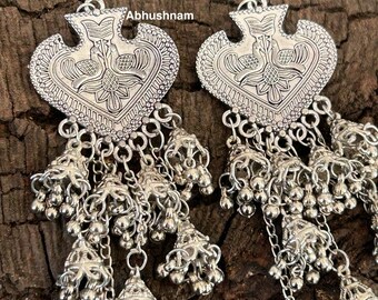 May include: Silver-toned earrings with a heart-shaped design and dangling bell charms. The earrings feature intricate patterns and the word "Abhushnam" is visible. The earrings are displayed on a textured, brown surface.
