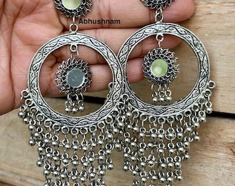 May include: Silver-toned chandelier earrings with a floral design and dangling beads. The earrings feature a circular design with a smaller floral accent in the center and a cluster of small beads hanging below. The earrings are held up by a person's hand.
