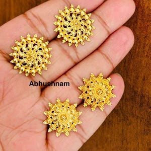 Pode incluir: São exibidos quatro brincos de botão dourados e ornamentados com um design de explosão solar. Cada brinco apresenta um padrão floral central rodeado por um anel de pequenas contas em relevo. A palavra "Abhushnam" é impressa na imagem.