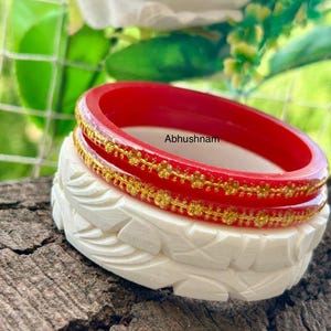 Op de afbeelding: Twee armbanden op elkaar. De bovenste armband is rood met gouden details, de onderste is wit met gesneden bladeren. De tekst "Abhushnam" is zichtbaar. De armbanden liggen op een houten oppervlak.