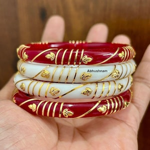 May include: Four bangles, alternating red and white, with gold leaf and line detailing. The red bangles have a shiny finish, and the white ones have a matte look. The text "Abhushnam" is visible on the white bangles.