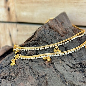 May include: Two gold anklets with dangling charms and small, clear gemstones. The anklets are displayed on a textured, dark brown surface, showcasing the jewelry's intricate design and sparkle. The anklets are a delicate accessory.