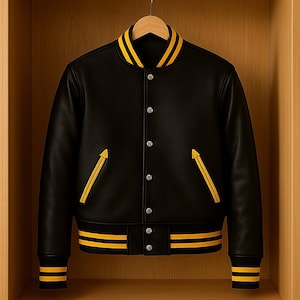 Puede incluir: Chaqueta universitaria negra con rayas amarillas en el cuello, los puños y el dobladillo. La chaqueta tiene detalles amarillos en los bolsillos y una botonadura frontal. Cuelga de una percha de madera en un armario de madera.