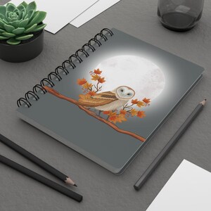 May include: A gray spiral bound notebook with a white barn owl perched on a branch with autumn leaves. The owl has a white face and brown and white feathers. A full moon is in the background.
