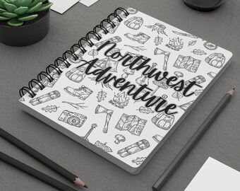 May include: A white spiral bound notebook with a black and white illustration of camping and outdoor adventure gear. The text "Northwest Adventure" is written in black cursive on the front cover.