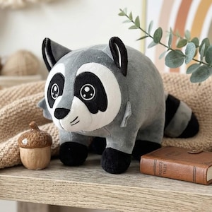 May include: A plush raccoon toy with grey fur, a white face mask, black eye patches, and a striped tail. The toy features embroidered eyes and a small black nose. It is positioned next to a small wooden acorn and a brown book titled Pudge's Tales.