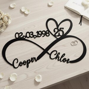 May include: A black metal infinity sign wall decoration featuring the names Cooper and Chloe in cursive script, the date 02.03.1998 with small heart accents, and a large heart shape containing the number 25, all displayed as a single cutout piece.