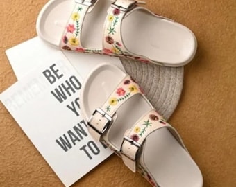 May include: A pair of white sandals with floral embroidery on the straps. The sandals have two adjustable buckles and a contoured footbed. A white card with the text "BE WHO YOU WANT TO" is next to the sandals.