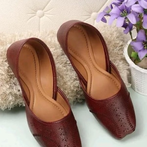 Puede incluir: Un par de bailarinas de cuero burdeos con punta y superficie texturizada. Los zapatos tienen un interior marrón claro y se exhiben sobre una superficie clara. Son un tipo de calzado.