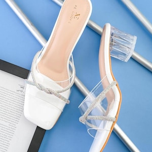 May include: A pair of white slide sandals featuring clear transparent straps, a square toe, and a block heel made of clear material. The straps are accented with a thin, silver-toned rhinestone-encrusted twist detail. The insole is white with the brand name XE LOOKS printed in black.