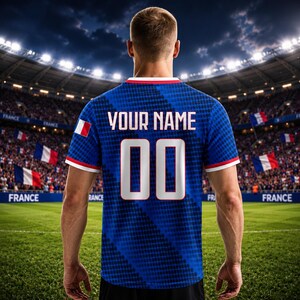 Op de afbeelding: Een blauw voetbalshirt met witte randen, met de Franse vlag op de mouw. De achterkant toont "YOUR NAME" boven het nummer "00" in wit. Het shirt wordt gedragen door een persoon op een groen veld in een stadion.