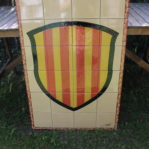 May include: A rectangular tile art piece featuring a shield design. The shield has vertical stripes in yellow, orange, and red, outlined in black and green. The border is beige with a decorative pattern.