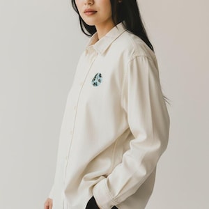 May include: Cream-colored button-down shirt with a small, round, embroidered design on the chest. The long-sleeved shirt has a classic collar and is paired with black pants. The embroidered design features shades of blue and green.