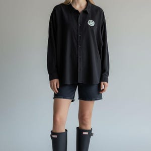 May include: Black long-sleeve button-down shirt with a small circular graphic on the chest. The shirt is paired with black denim shorts and black rubber boots. The outfit is casual and suitable for various occasions.