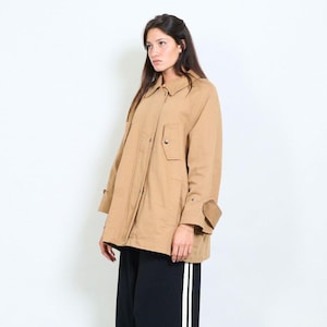 May include: A tan jacket with a collared neckline, button fastenings, and a small pocket. The jacket has long sleeves with buttoned cuffs. The person is wearing black trousers with white stripes.