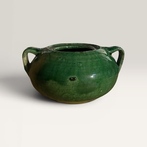 Puede incluir: Una olla redonda de cerámica verde con dos asas. La olla tiene una abertura ancha y una superficie texturizada. El color es un verde vibrante y brillante, con algunas áreas más oscuras. La olla es probablemente para uso decorativo o funcional.
