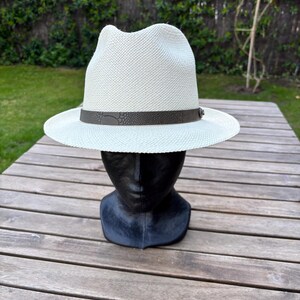May include: A white fedora hat with a dark brown band, displayed on a black mannequin head. The hat has a woven texture and a classic brim. The background features a wooden table and green foliage.