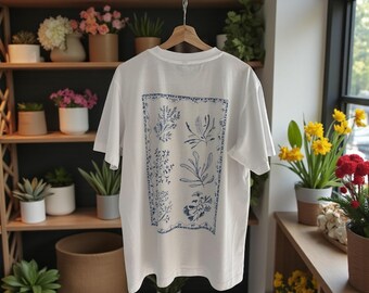 Könnte beinhalten: Weißes T-Shirt mit blauem botanischem Druck auf dem Rücken. Das Design zeigt verschiedene Pflanzenillustrationen in einem rechteckigen Rahmen. Das T-Shirt hängt an einem Holzbügel vor dem Hintergrund von Pflanzen und Regalen.