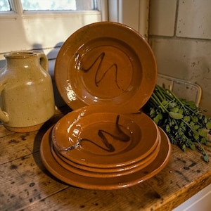 Puede incluir: Un conjunto de platos de cerámica de color terracota con un diseño de garabatos marrón oscuro. Una jarra a juego es visible a la izquierda. Los platos están sobre una superficie de madera rústica.