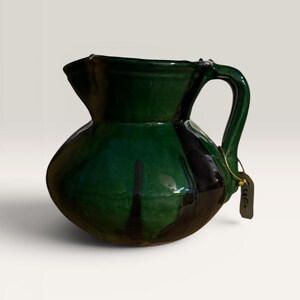 Può includere: Una brocca in ceramica verde con corpo arrotondato e manico curvo. La brocca ha un beccuccio per versare e una glassa marrone scuro che cola lungo il lato. Una piccola etichetta è attaccata al manico.