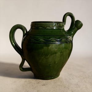 May include: A handcrafted, green ceramic pitcher with a spout and two handles. The pitcher has a glossy finish and decorative wavy lines around the top. The design is a classic, rustic style.