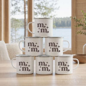 May include: A stack of six white ceramic mugs with a silver rim, arranged in a pyramid. Each mug features a dark brown graphic design consisting of the lowercase letter m repeated twice, accompanied by four-pointed star icons on the front.