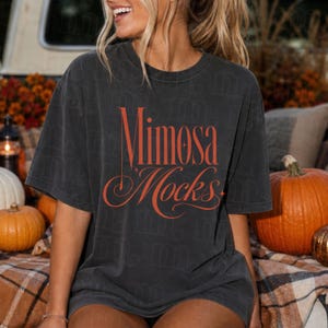 Puede incluir: Una camiseta de manga corta gris carbón con el texto Mimosa Mocks en una fuente serif naranja estilizada. La prenda tiene cuello redondo y un corte relajado, con un sutil patrón de marca de agua repetido visible en la tela.