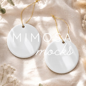 Può includere: Due ornamenti circolari in ceramica bianca con superfici lisce e lucide. Ogni ornamento presenta un piccolo foro nella parte superiore con un sottile cordino dorato metallizzato per appenderlo. Il testo "MIMOSA mocks" è sovrapposto in bianco.
