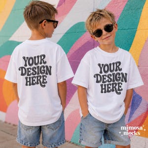 Może przedstawiać: Biały t-shirt z krótkim rękawem z czarnym napisem YOUR DESIGN HERE nadrukowanym z przodu i z tyłu. Koszulka jest noszona z niebieskimi szortami dżinsowymi i czarnymi okularami przeciwsłonecznymi. Projekt wykorzystuje pogrubioną, zaokrągloną czcionkę.