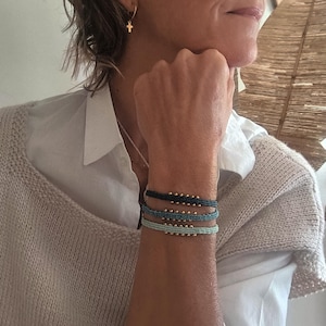 Peut inclure: Trois bracelets de perles bleues, bleu marine et bleu clair ornent un poignet. Chaque bracelet est orné de petites perles dorées et d'un motif tissé. Les bracelets créent un effet superposé. La personne porte une chemise blanche et un pull beige.