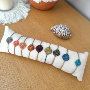 Peut inclure: Une collection de bracelets en forme de feuilles perlées de différentes couleurs, présentés sur un coussin blanc. Les bracelets ont des chaînes dorées. Un coquillage et des objets décoratifs sont en arrière-plan.