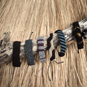 Peut inclure: Plusieurs bracelets en macramé faits à la main, de couleurs et de motifs variés, dont noir, bleu, gris et beige. Les bracelets sont exposés sur une branche de bois vieilli, sur fond d'herbes sèches.