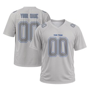 May include: A light gray short-sleeved sports jersey with a V-neck collar. The front features the text "YOUR TEAM" above the number "00" in gray with blue outlines. The back displays "YOUR NAME" above the number "00" in the same color scheme.
