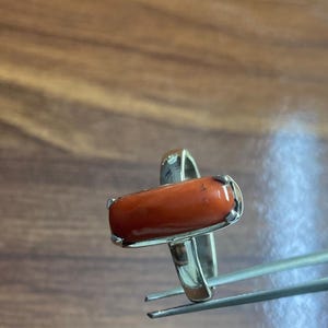 May include: A silver ring featuring a vibrant orange-red, oblong gemstone. The gemstone is set in a silver bezel, and the ring is held by tweezers against a wooden background. The ring's design is simple yet elegant.