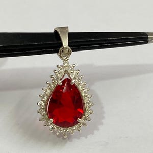 Pode incluir: Um pingente de prata com uma grande pedra preciosa vermelha em forma de gota. A pedra preciosa é cercada por pedras menores e transparentes. O pingente é suspenso por uma argola de prata. A joia é exibida em um fundo branco.
