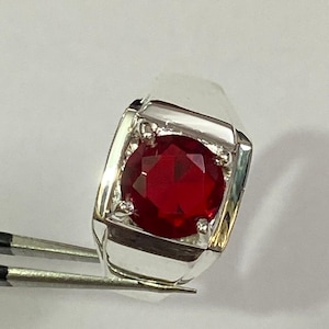 Może przedstawiać: Srebrny pierścionek z dużą, okrągłą, głęboko czerwoną kamieniem szlachetnym. Kamień jest osadzony w kwadratowej oprawie z polerowaną srebrną obrączką. Pierścionek jest trzymany przez pęsetę na białym tle.