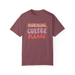 Może przedstawiać: Bordowa koszulka z napisem "MORNING, COFFEE PLEASE" w stylu retro. Tekst jest warstwowy w odcieniach pomarańczu, różu i czerwieni. Koszulka ma krótkie rękawy i okrągły dekolt.