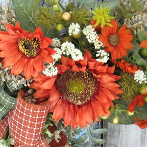 May include: A decorative wreath featuring large orange sunflowers with dark centers, small white filler flowers, green foliage, and textured berries. The arrangement is accented with a burnt orange ribbon featuring a white grid pattern.