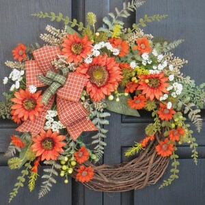 May include: A decorative wreath featuring orange sunflowers, small white flowers, and green fern foliage arranged on a circular brown twig base. An orange and white checkered ribbon with a green accent bow is tied to the left side of the floral arrangement.