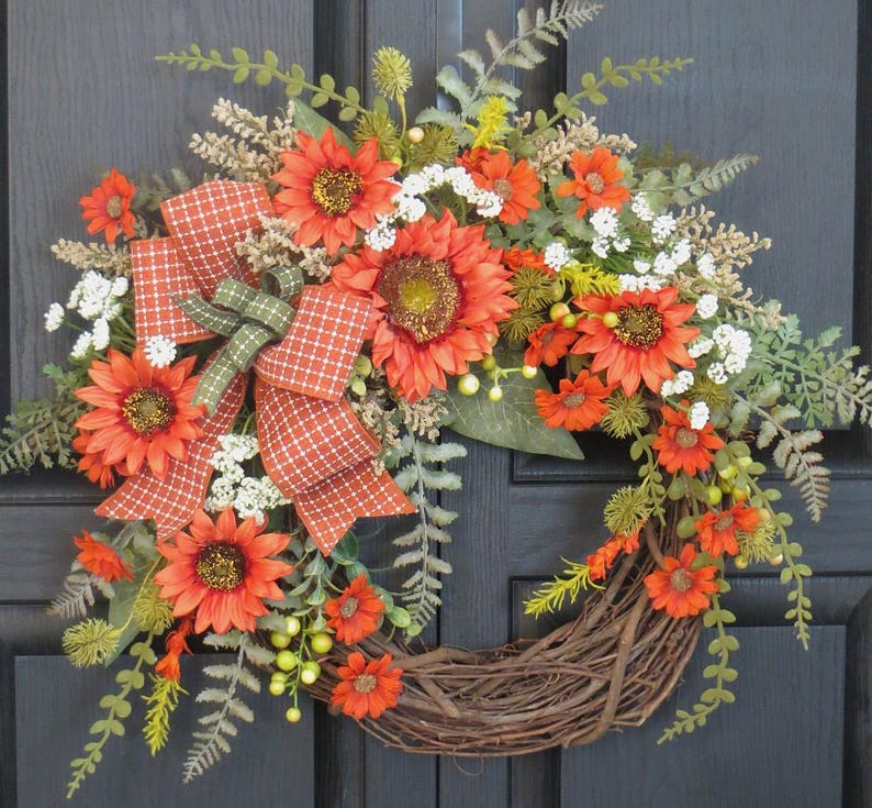 May include: A decorative wreath featuring orange sunflowers, small white flowers, and green fern foliage arranged on a circular brown twig base. An orange and white checkered ribbon with a green accent bow is tied to the left side of the floral arrangement.