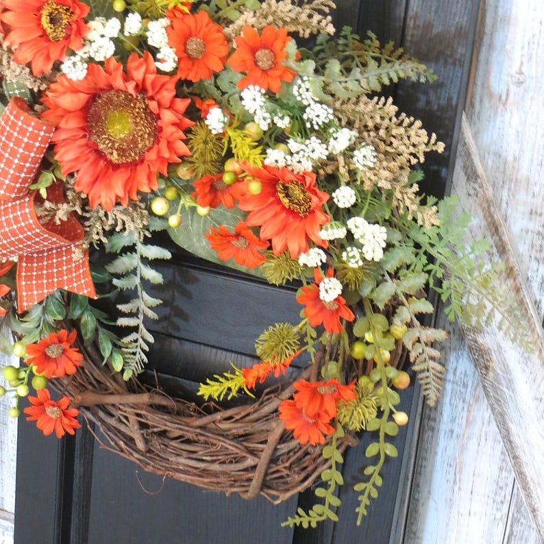 May include: A decorative wreath featuring vibrant orange sunflowers and smaller orange daisies, accented with white baby's breath, green foliage, and textured fern sprigs. An orange and white checkered ribbon bow is attached to the side.